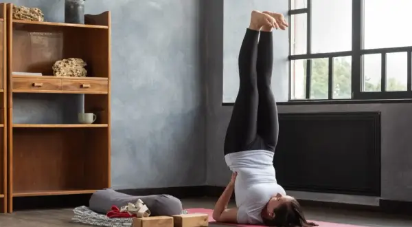 Frau praktiziert Iyengar Yoga in der Umkehrhaltung (Sarvangasana) auf einer rosa Matte, unterstützt von Holzblöcken.