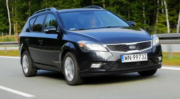Czarny Kia Ceed 1. kombi jedzie po drodze, otoczony zielenią.