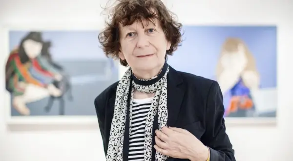 Ewa Kuryluk, artystka o burzy brązowych włosów, w czarnej marynarce i pasiastym szalu, pozuje na tle swoich obrazów.