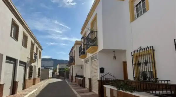 Calle soleada con casas blancas y detalles amarillos en Celin, Almería. Balcones de hierro forjado y un cielo azul con nubes.
