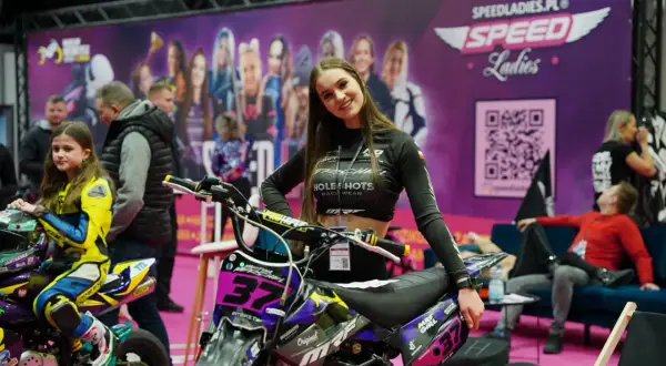 Uśmiechnięta dziewczyna na tle banera z napisem "Warsaw Motorcycle Show" i motocyklem. Obok młoda zawodniczka w żółtym kombinezonie.