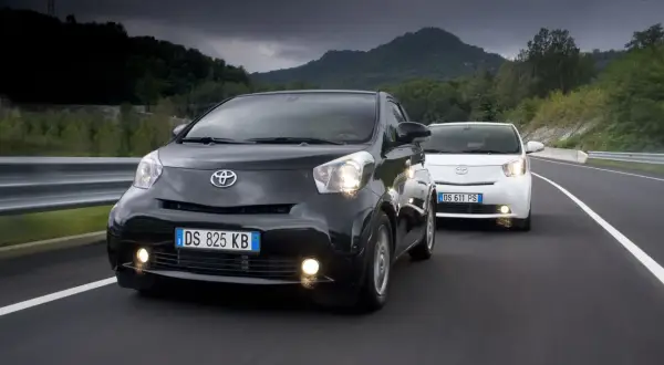 Dwa samochody Toyota iQ, czarny i biały, jadące po drodze w malowniczym, górzystym krajobrazie.