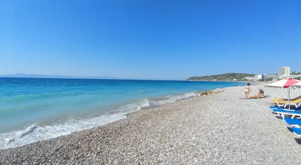 Słoneczna plaża w Ixia, Rodos, z turkusowym morzem i kamienistym brzegiem. Dwie osoby odpoczywają pod parasolem.