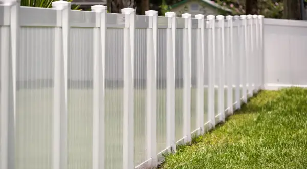A newly completed vinyl fence installation, featuring white posts and translucent panels, lines a lush green lawn.