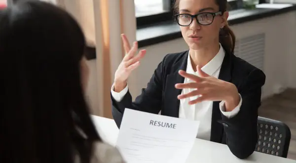 Kobieta w okularach i czarnej marynarce prowadzi rozmowę kwalifikacyjną, gestykulując rękami nad dokumentem "RESUME".
