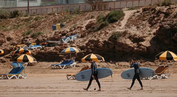 Deux surfeurs marchent sur la plage de Tamraght, Maroc, avec leurs planches sous le bras. Des parasols et des chaises longues sont installés sur le sable.