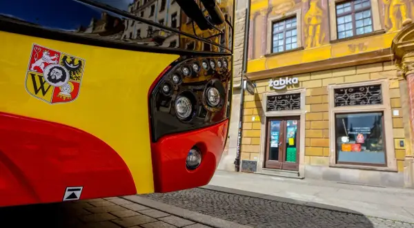 Żółto-czerwony autobus 143 Kraków z herbem miasta na błotniku, zaparkowany przed kamienicą z malowidłami i sklepem Żabka.