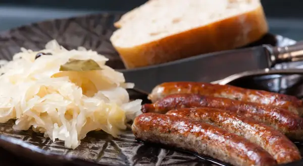 Herzhaftes Sauerkraut mit vier goldbraunen Würsten und einem Stück Brot auf einem dunklen Teller.