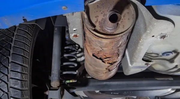 Vue du dessous d'une voiture bleue, montrant une roue, un ressort de suspension et un silencieux rouillé et endommagé.