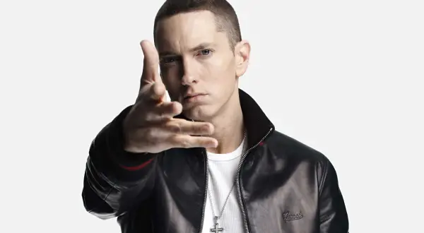 Eminem w czarnej kurtce i białej koszulce, gestykuluje ręką.