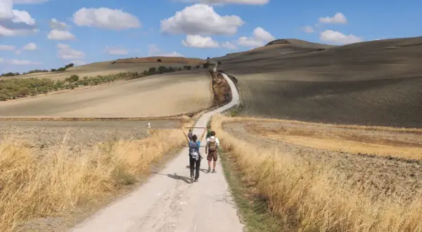 Due pellegrini camminano lungo la Via Francigena, immersi in un paesaggio collinare toscano sotto un cielo azzurro con nuvole bianche.