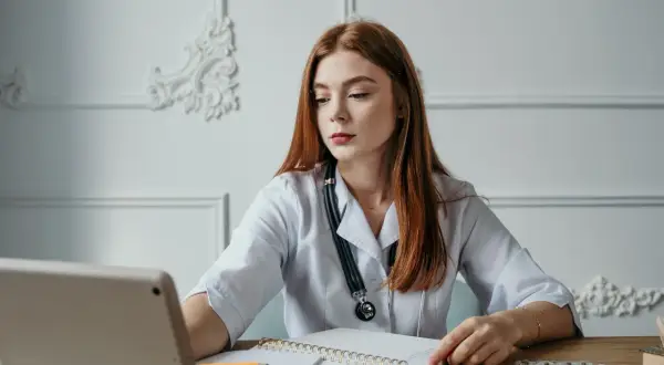 Młoda lekarka z rudymi włosami, w białym fartuchu, z notatnikiem i tabletami, szuka informacji o hemoroidach jaki lekarz.
