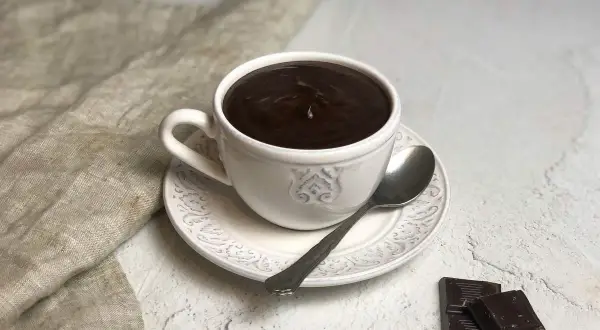 Una tazza di cioccolato caldo fumante, con cucchiaino e tavolette di cioccolato fondente accanto.