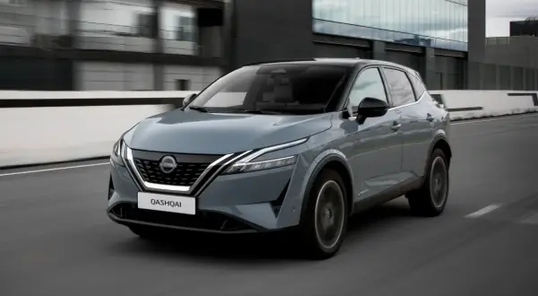 Grå Nissan Qashqai kör på en väg, en del av ett nissan qashqai test. Moderna byggnader syns i bakgrunden.