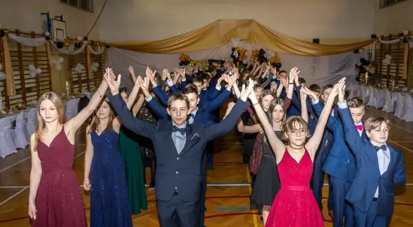 Młodzież w eleganckich strojach, dziewczęta w sukniach, chłopcy w garniturach, tańczą na szkolnym balu. To ich pierwszy komers, pełen radości i wspólnej zabawy.