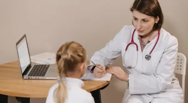 Lekarka w białym fartuchu rozmawia z dzieckiem przy biurku z laptopem.