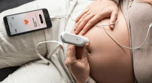 Manos de mujer embarazada usando un doppler fetal para escuchar el latido del bebé. La pantalla del móvil muestra "Escuchando latidos".
