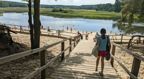 Dziewczynka schodzi drewnianymi schodami na piaszczystą plażę nad jeziorem. W oddali widać ludzi kąpiących się i odpoczywających. Stawy echo zwierzyniec to idealne miejsce na letni relaks.