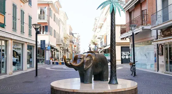 Scultura di elefante e palma sul centro passeggiata San Benedetto del Tronto, con negozi e palazzi colorati sullo sfondo.