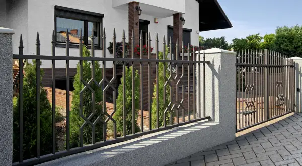 Elegancka brama przesuwna czy dwuskrzydłowa? Metalowe ogrodzenie z ozdobnymi elementami i cyprysami przed domem.