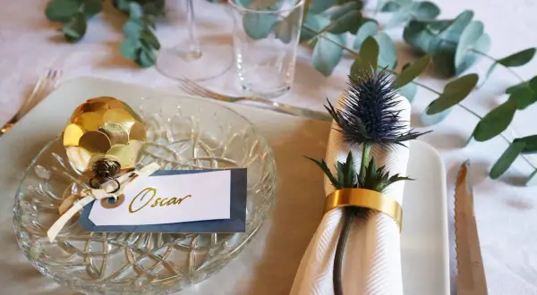 Elegant nyårsdukning med gulddekorationer och en namnskylt för Oscar. En gyllene kotte pryder tallriken, medan en blå tistel med guldfärgad servettring skapar en vacker detalj.