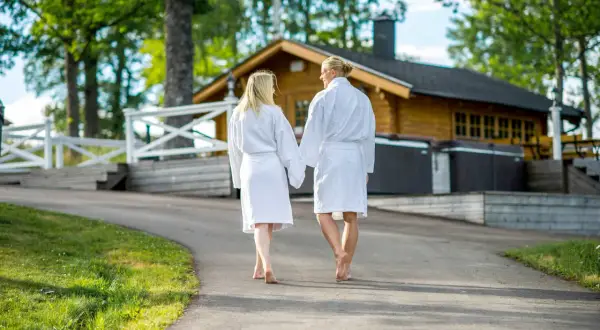 Ett par i badrockar går hand i hand mot en timrad stuga med utomhuspool. Kanske på väg till en avkopplande stund vid hvide sande.