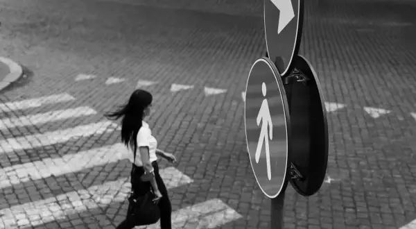 Una donna attraversa la strada su un selciato, seguendo le indicazioni di un segnale pedonale. Fotografia di strada.