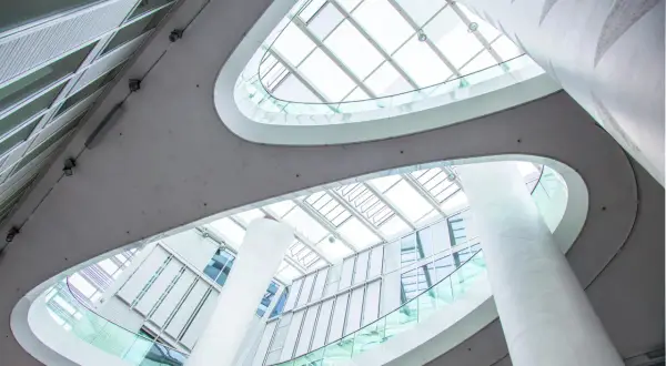 Nowoczesne wnętrze z białymi kolumnami i szklanymi balustradami, przez które wpada obfite światło dzienne.