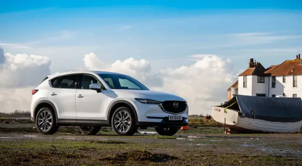 Vit Mazda CX-5 parkerad vid en båt på en lerig strand, med hus och moln i bakgrunden.