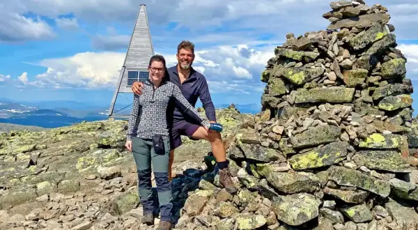 Två glada vandrare vid en stenröse på en bergstopp i Sverige. En modern, pyramidformad byggnad med solpaneler syns i bakgrunden. Perfekt för att utforska Sveriges vandringsleder.