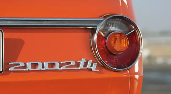 Detalle de la parte trasera de un BMW 2002 Tii naranja, con su icónico emblema cromado y una luz trasera circular.