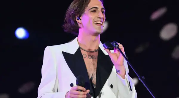 Damiano David w białej marynarce z czarnym kołnierzem śpiewa na koncercie, trzymając mikrofon.
