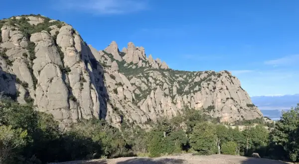 Imponentes rocas de Montserrat bajo un cielo azul, con vegetación verde y la creu de Sant Miquel en la distancia.