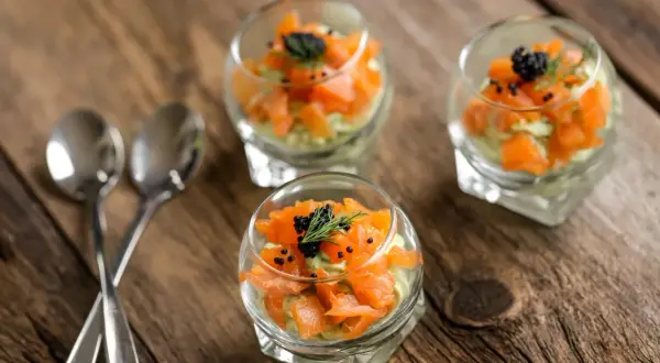Trois verrines de saumon, avocat et caviar, garnies d'aneth, sur une table en bois. Deux cuillères argentées sont posées à gauche.
