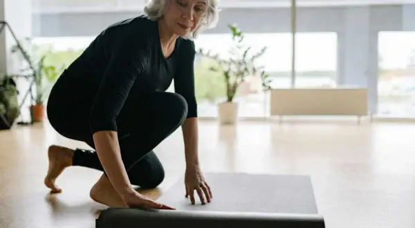 Une femme prépare son tapis pour faire du sport chez soi. Elle est à genoux, déroulant le tapis gris sur le sol en bois.