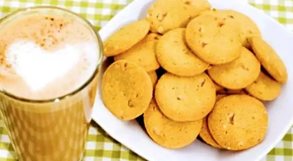 A glass of coffee with a heart-shaped foam art next to a plate of golden brown, round cookies.