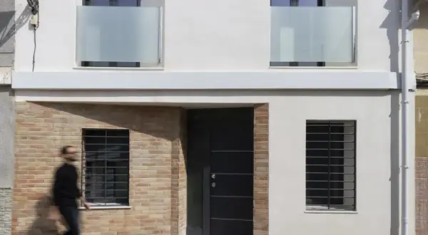 Fachada de casa con ladrillo visto y pared blanca. Una puerta oscura y ventanas con rejas. Ideas para como construir una casa barata en España.