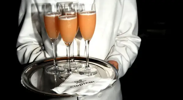 Kellner serviert Bellinis auf einem Silbertablett. So trinkt man Bellini: ein erfrischender Cocktail aus Prosecco und Pfirsichpüree.