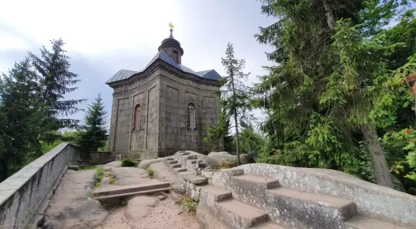 Kamienna kaplica na skale, otoczona drzewami. Broumowskie ściany budowli wznoszą się ku niebu, a kamienne schody prowadzą do jej wnętrza.