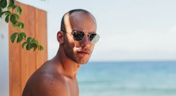 Uomo con capelli cortissimi, occhiali da sole e sfondo mare.