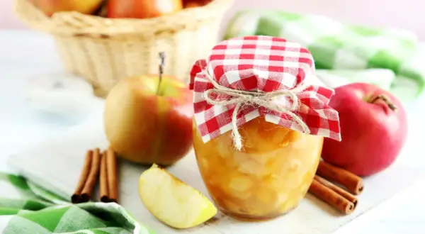 Frisch eingekochtes Apfelkompott im Glas, dekoriert mit kariertem Stoff und Zimtstangen. Äpfel im Korb im Hintergrund.