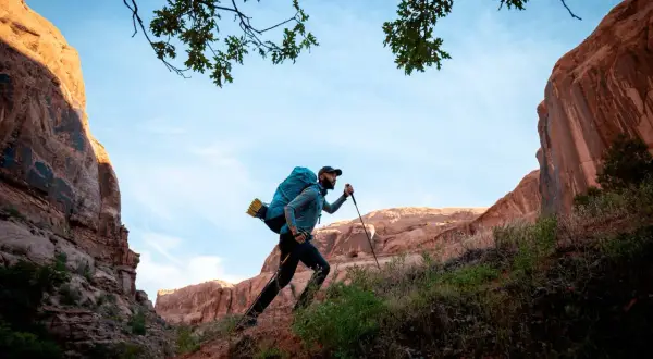 Hombre con mochila y bastones de senderismo sube una ladera rocosa bajo un cielo azul.