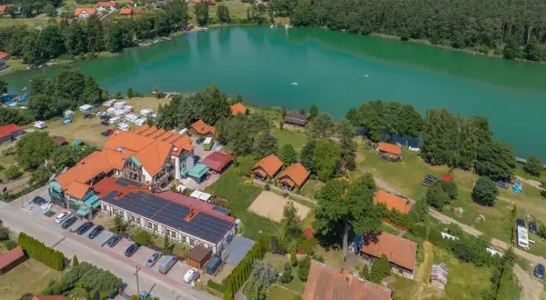 Widok z lotu ptaka na ośrodek wypoczynkowy nad turkusowym jeziorem. Widać budynki, domki, pole namiotowe, kampery i plażę. To idealne miejsce na wypoczynek, niczym złota łania wiartel.