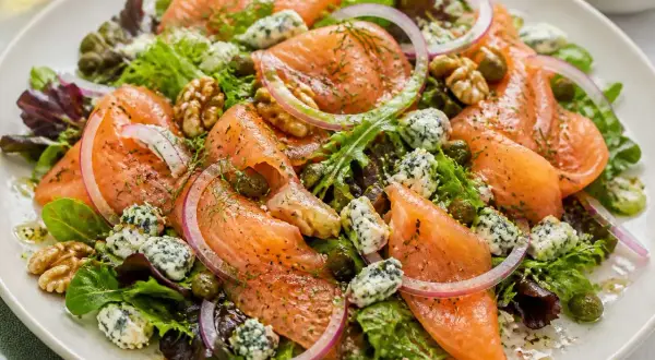 Salade colorée avec saumon fumé, fromage bleu émietté, noix, câpres et oignons rouges sur un lit de verdure.