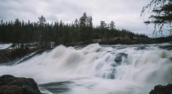 Vilt och mäktigt vattenfall i Norrland, med tät barrskog som bakgrund och en dramatisk, grå himmel.