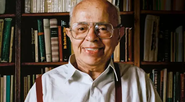Uśmiechnięty Stanisław Lem, autor powieści Solaris, siedzi przed regałami pełnymi książek.