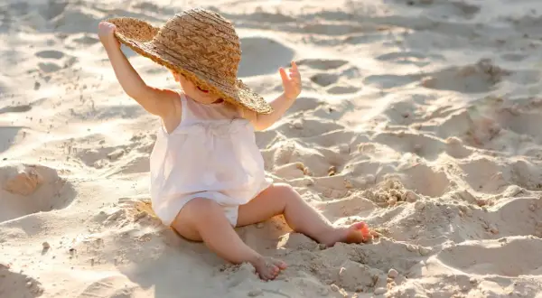 Liten bebis på stranden med solskyddsfaktor, leker med en stor stråhatt.