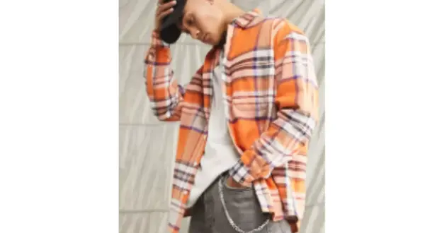 Uomo con camicia in flanella a quadri arancioni, jeans grigi e cappellino nero.