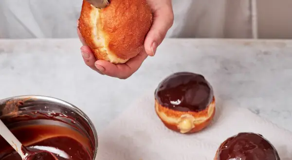 Eine Hand füllt einen Boston Cream Donut mit cremiger Füllung. Daneben liegen bereits glasierte Donuts.