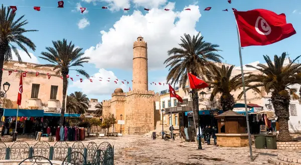 Placez-vous ici pour savoir où aller en Tunisie : une place animée avec des drapeaux tunisiens, des palmiers et une tour historique.